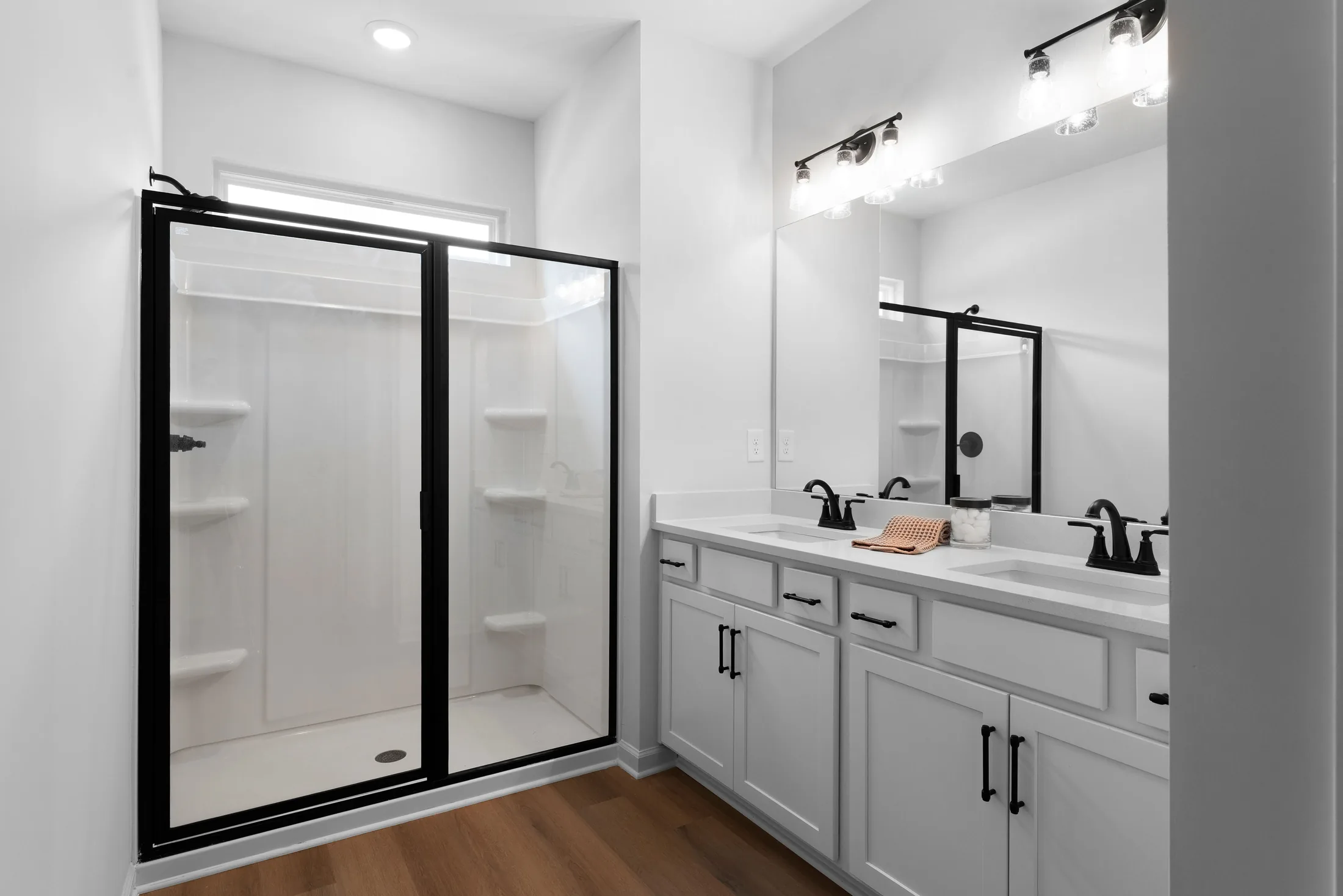 Modern bathroom with a glass shower enclosure, double sink vanity with white cabinets, black fixtures, mirror, and wooden flooring.
