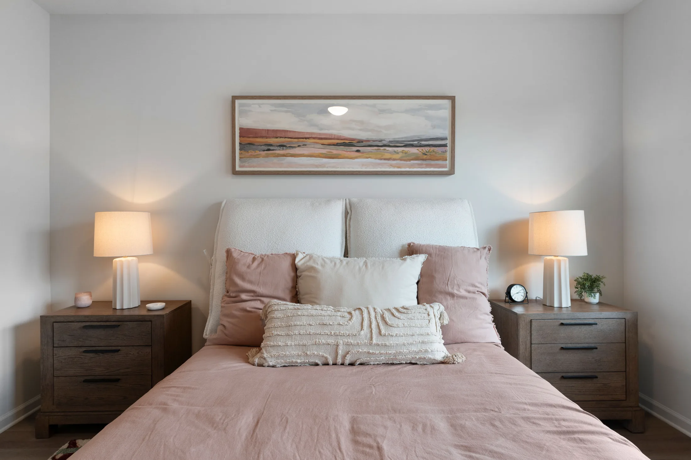 A neatly made bed with pink and white bedding is centered between two dark wood nightstands, each holding a lamp. A landscape painting hangs above the bed.