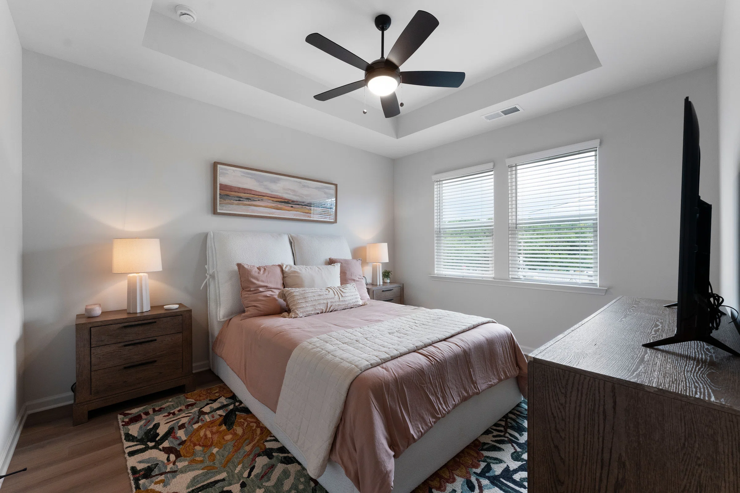 A bedroom with a bed, two nightstands with lamps, a ceiling fan, a dresser with a TV, a window with blinds, and a landscape painting above the bed.