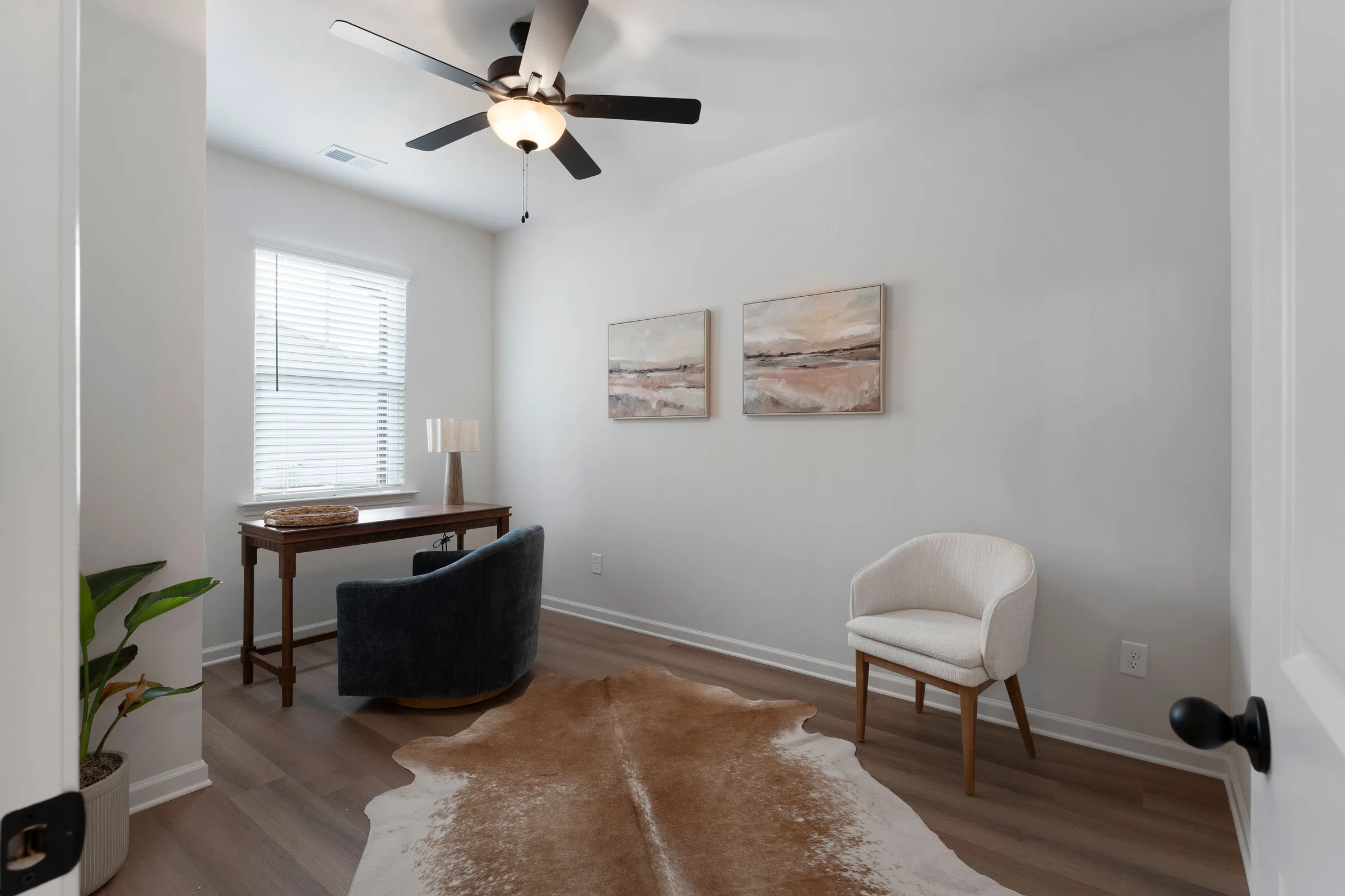 Small office with a wood desk, gray chair, white armchair, cowhide rug, two wall art pieces, and a ceiling fan with lights. A plant sits in the corner by the window with white blinds.