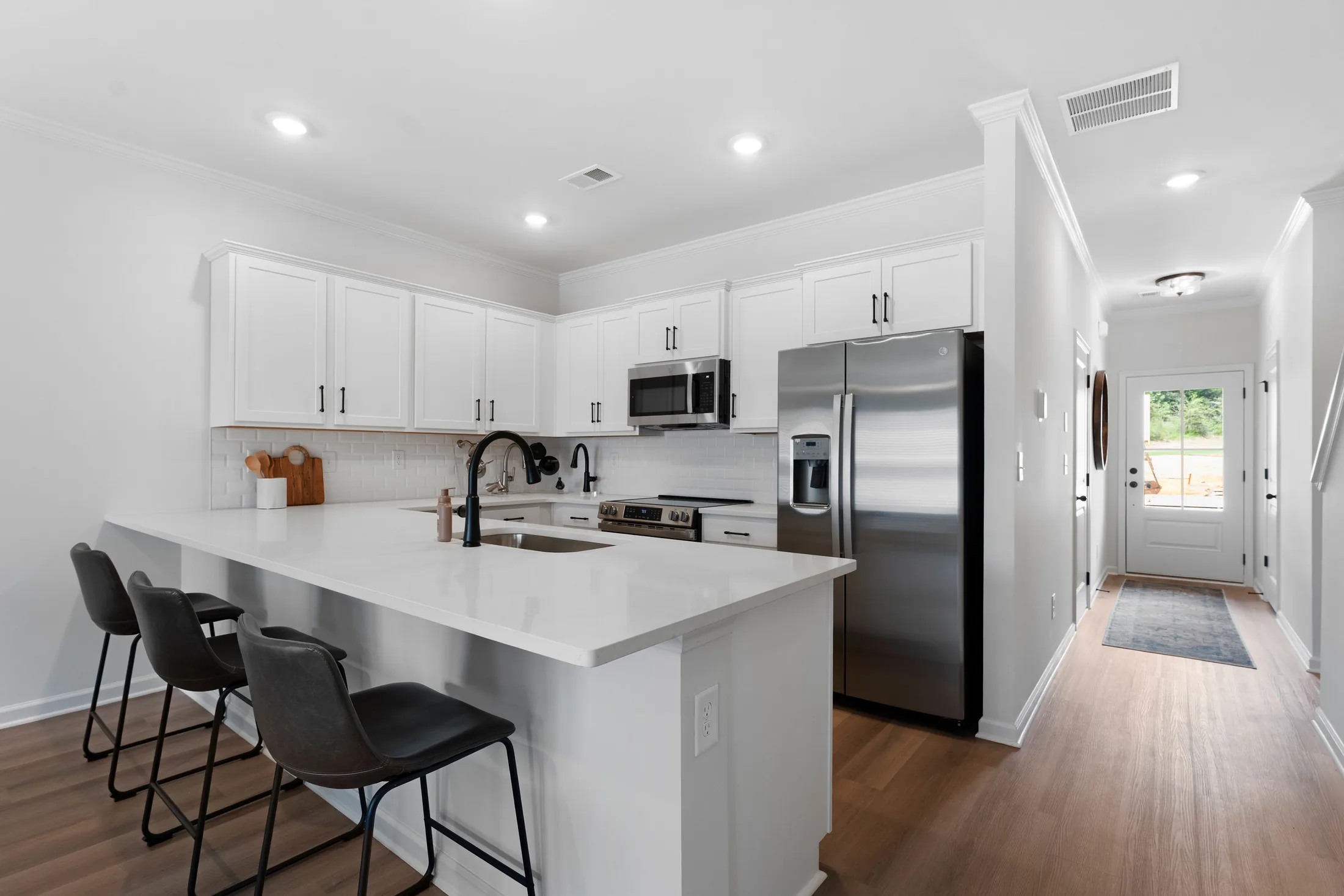Modern home kitchen with white cabinets, stainless steel appliances, a sink island with three black chairs, and light wood flooring. A hallway with a rug leads to a door with windows at the back, creating a bright and inviting space perfect for displaying your favorite memories from Picture Life Gallery.
