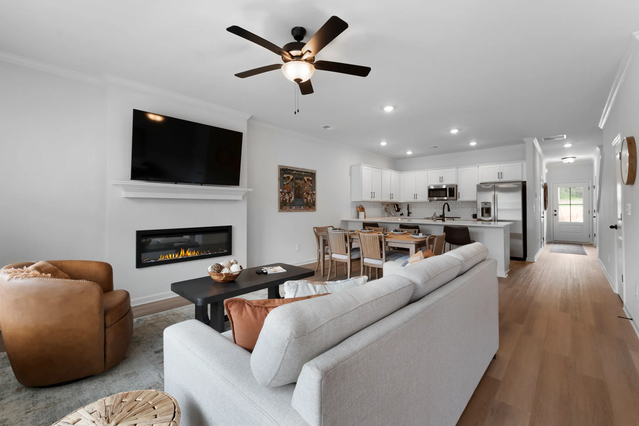Open-concept living room and kitchen in Home 2 with a gray sofa, round chairs, wall-mounted TV, fireplace, dining table, and modern white cabinetry under recessed lighting—perfectly designed to showcase your favorite memories from the Picture Life Gallery on The Boulevard.