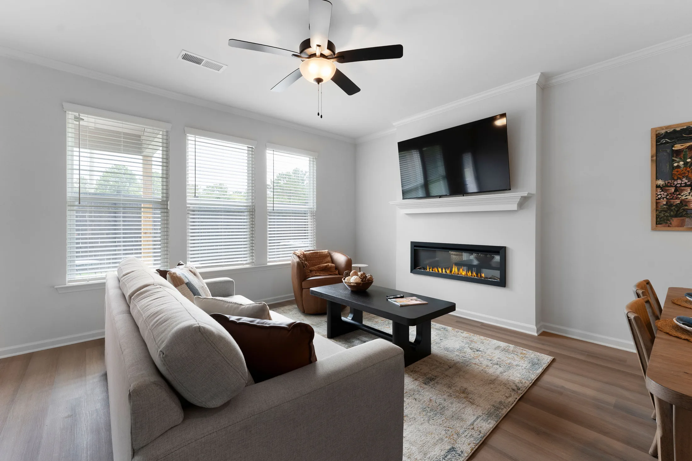 Modern living room with a beige sofa, black coffee table, armchair, wall-mounted TV, fireplace, large windows, and light wood flooring.