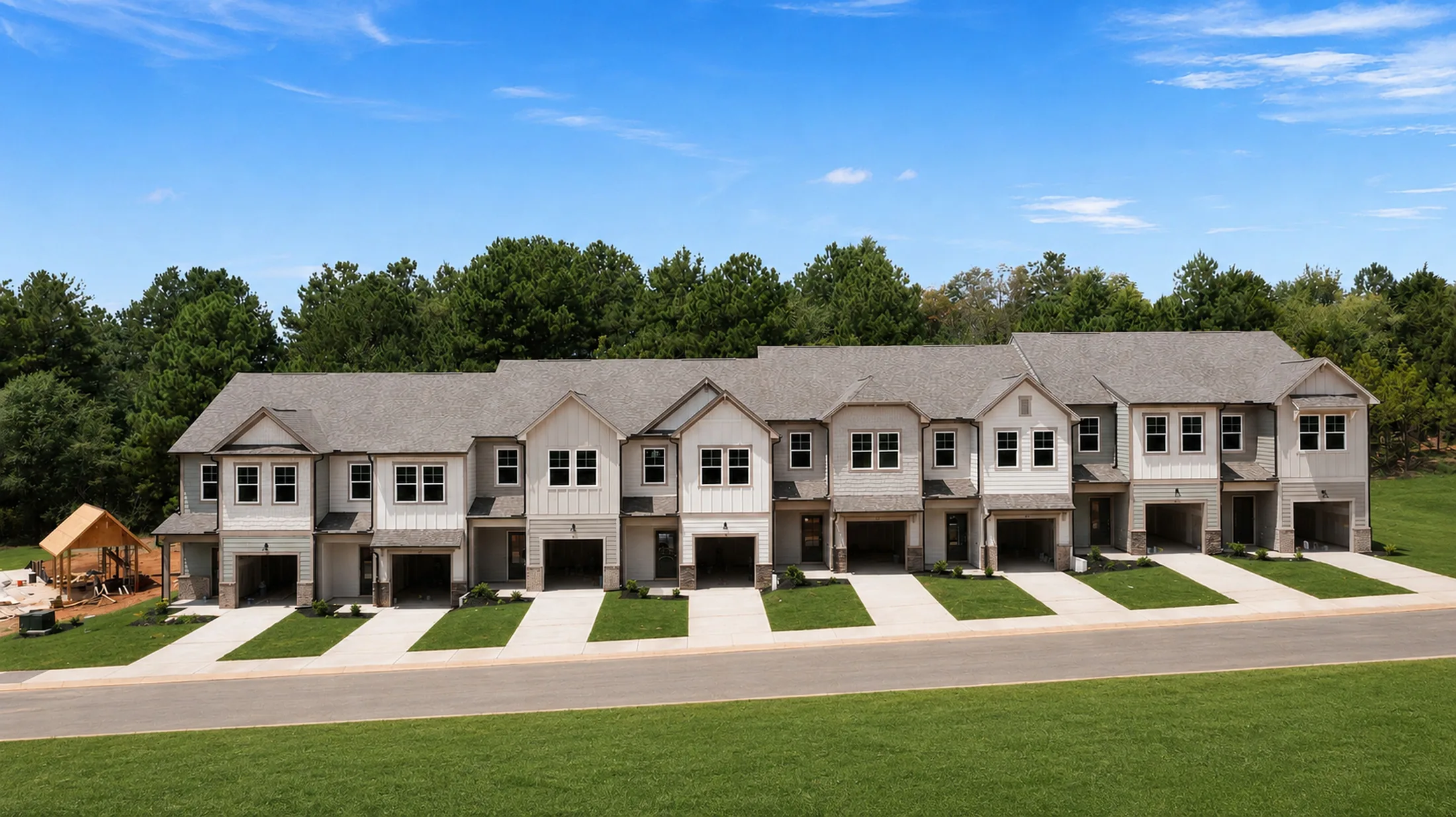 Row of modern two-story townhouses with garages, set on neatly mowed lawns, with a wooded area and blue sky in the background.