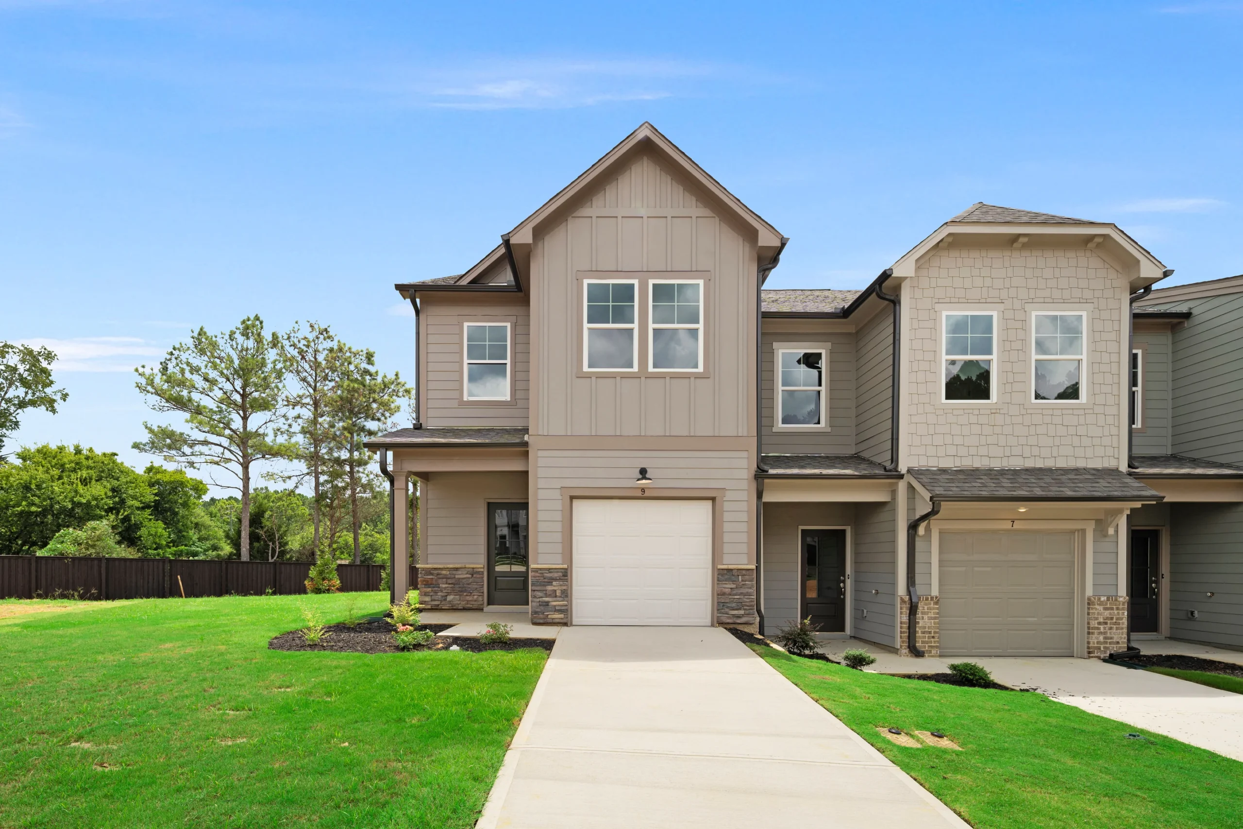 Modern two-story townhouse with a single-car garage, beige siding, multiple windows, and a well-kept lawn, set against a clear blue sky.