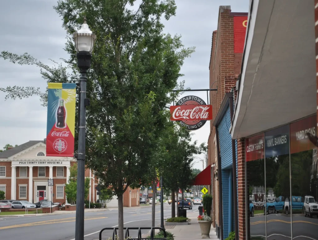 A small town street in Cedartown shows a Coca-Cola sign, a lamp post, and a courthouse in the background under a cloudy sky, capturing community life.