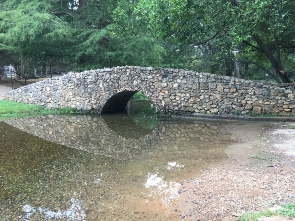 A small stone footbridge arches over a shallow creek in a wooded park area, with trees and greenery in the background.
