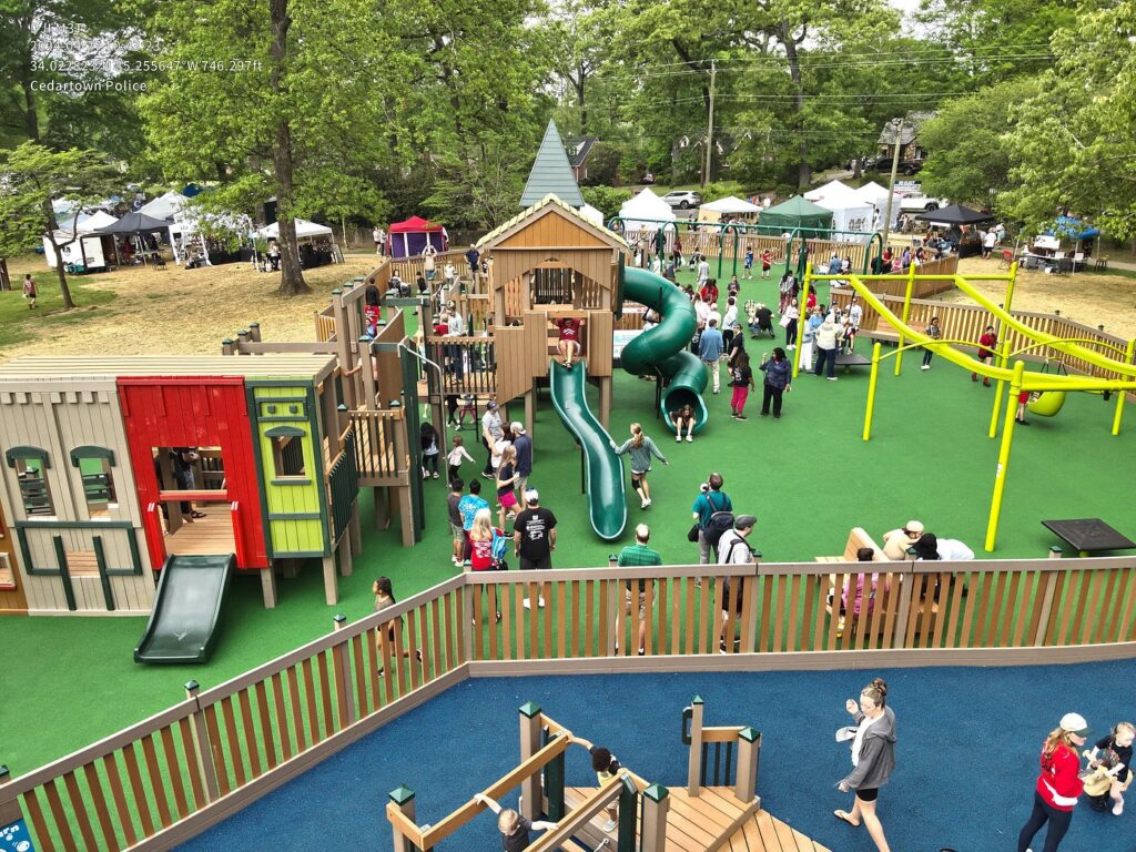 Cedartown community playground with play structures, a slide, green turf, and tents and trees in the background.