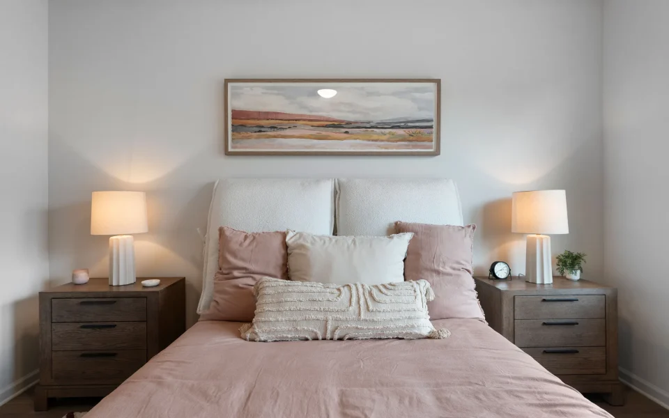 A bed with a white headboard and pink bedding, flanked by two wooden nightstands with lamps and a landscape painting centered above.