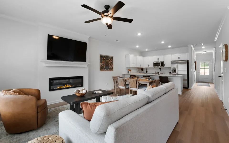 An open-concept living area with a fireplace, mounted television, light-colored sofa, leather armchair, and a kitchen with a dining table in the background.