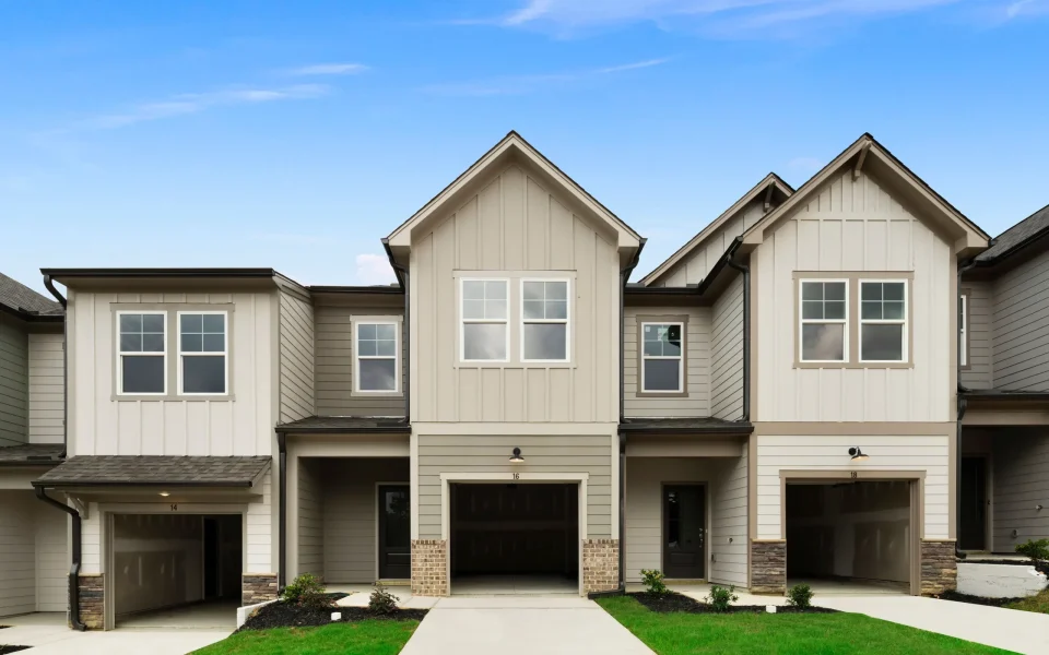 Row of modern beige townhouses with white trim, attached garages, neatly trimmed grass lawns, and a blue sky in the background.